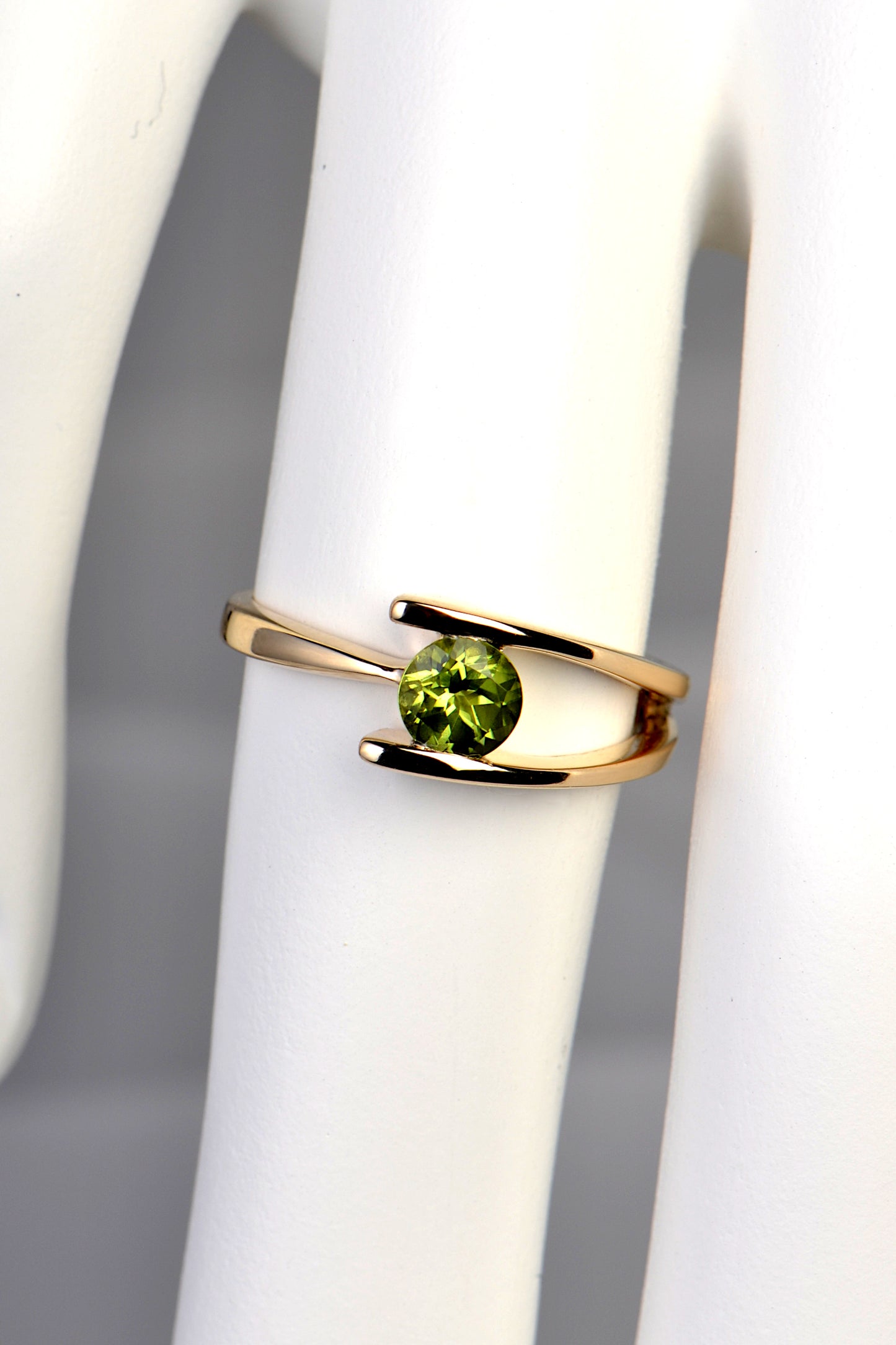 peridot and yellow gold designer ring on a finger. It has a round peridot in the middle with two bands on the right side that join into one at the back and appear on the left side of the stone as one band