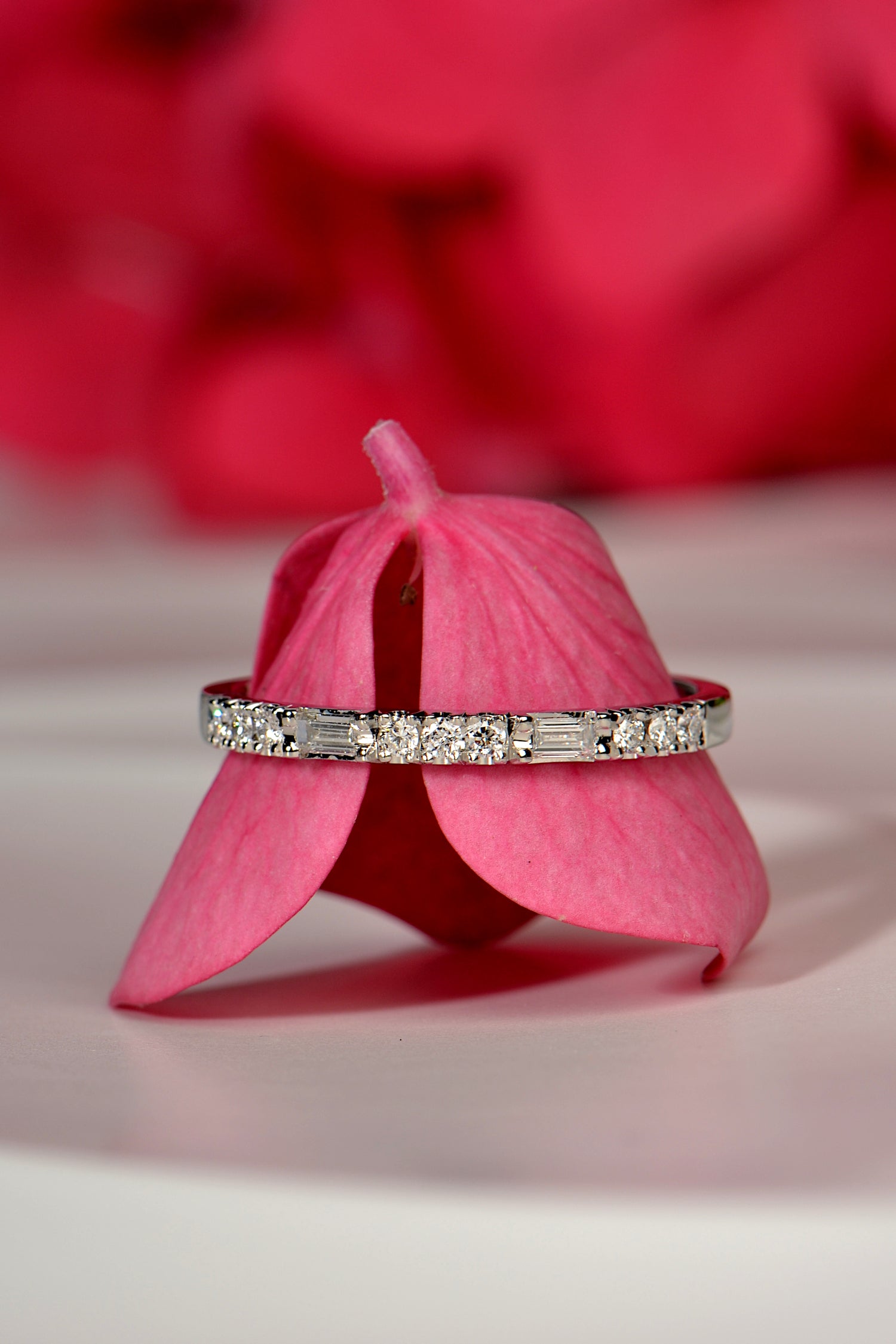 modern narrow platinum diamond set wedding ring with baguette and round diamonds in a dot dot dot dash design photographed on a cerise pink flower petal