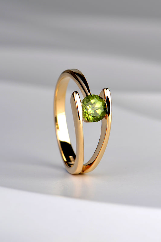 modern asymmetric ring in yellow gold that has two bands on one side that merge into one on the other with a round peridot gemstone that is olive green in colour