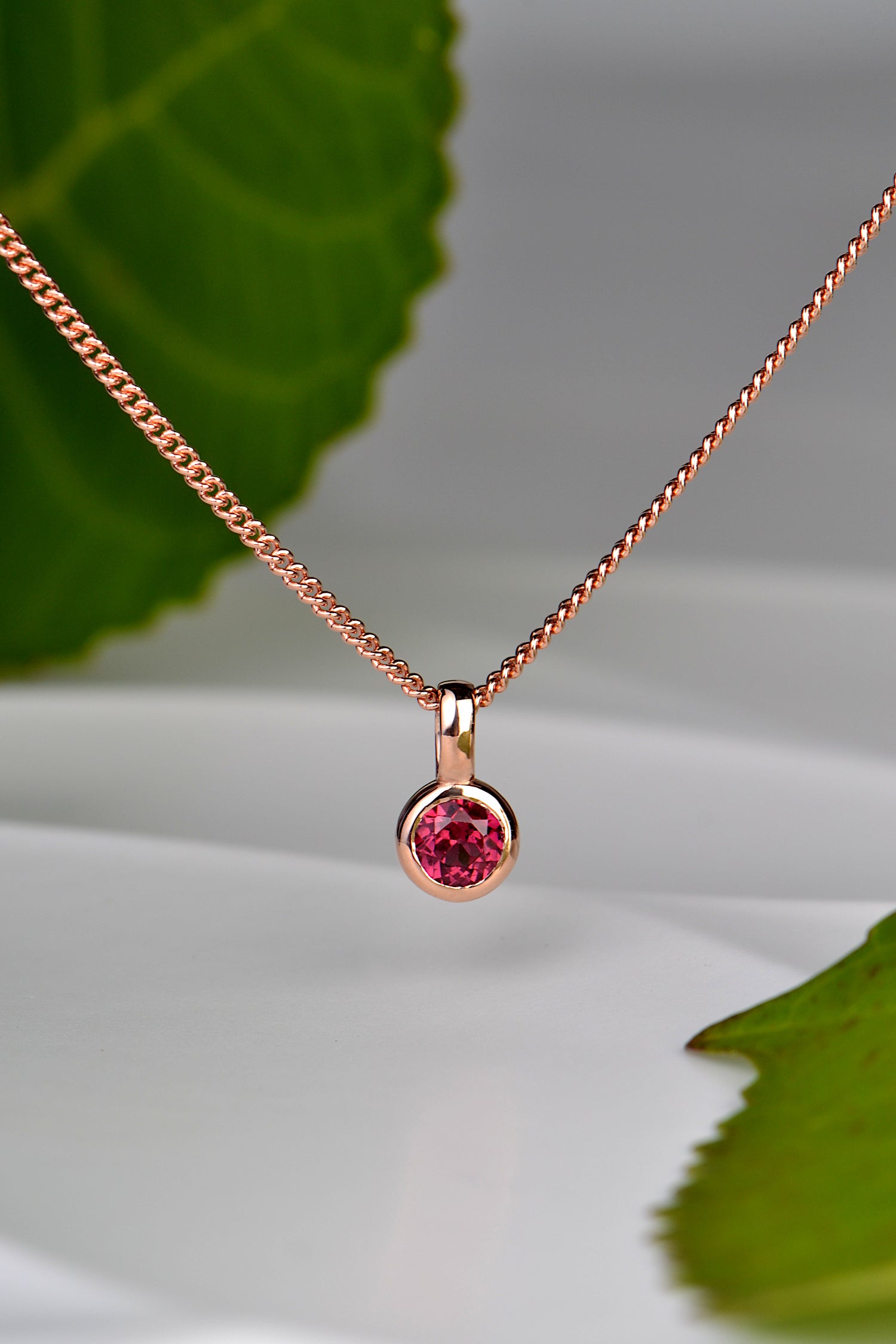 cerise pink tourmaline in 9ct rose gold pendant on 45cm long curb chain with two green leaves in the background