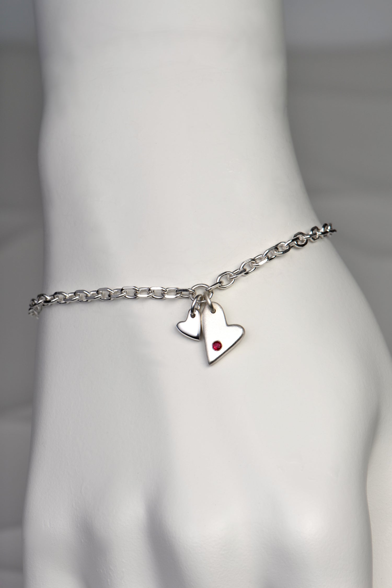 handmade silver heart bracelet by Christine Sadler. Two hearts hang at the front, one larger than the other. The larger heart is matt finished and set with a ruby gemstone